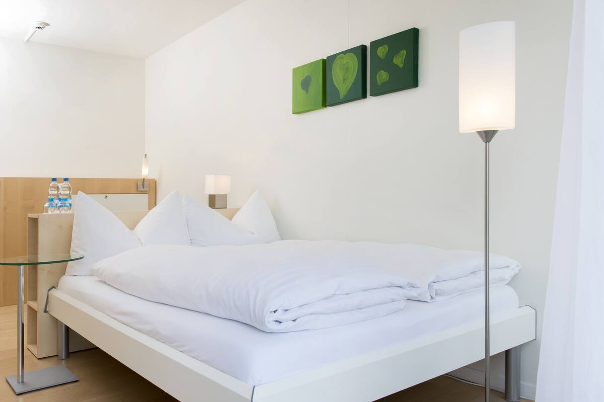 A neatly made bed with white linens, a glass side table, a bedside lamp, and a wall-mounted artwork.