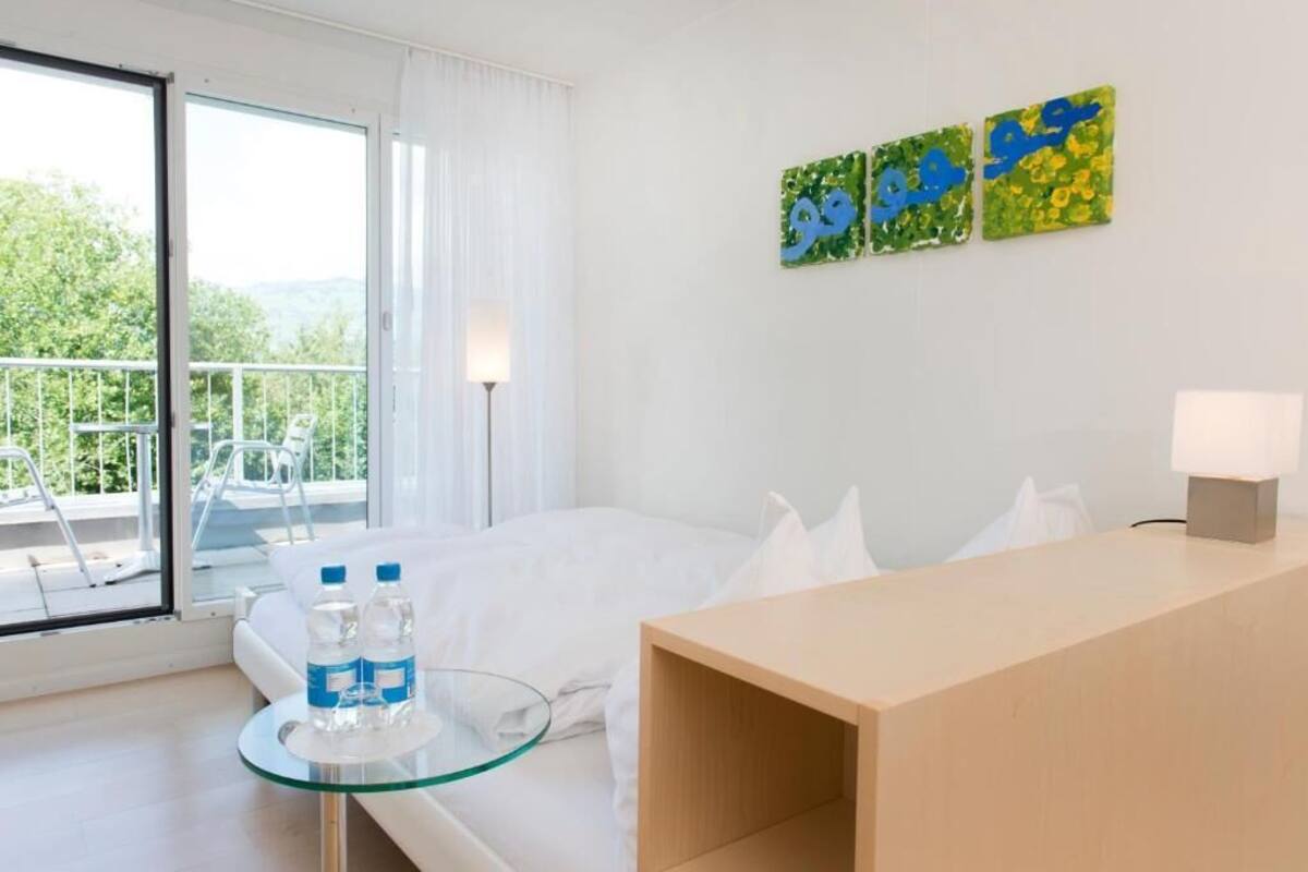 A hotel room with a bed, a glass table with water bottles, a wooden desk, and a balcony with a chair.
