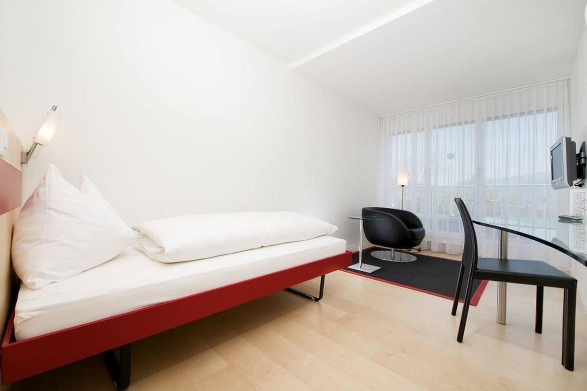 A modern hotel room with a red-framed bed, a black chair, and a TV mounted on the wall.