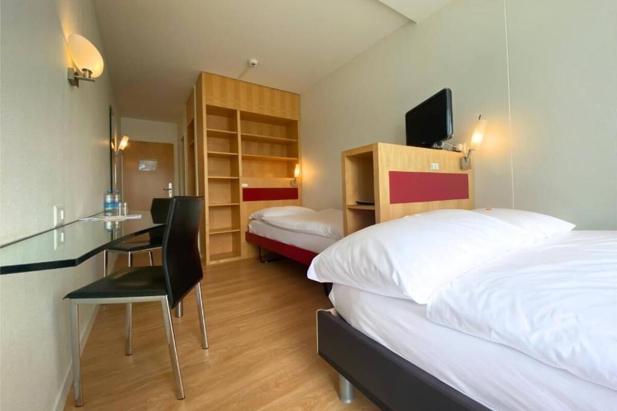A hotel room with a bed, a desk with a chair, a TV, and a wooden shelving unit.