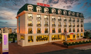 Um hotel com vários andares, um grande letreiro "ANEMON", janelas amplas e varandas com floreiras.