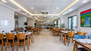 Un comedor amplio con múltiples mesas y sillas, una cocina central, y un ventanal con vistas a la ciudad.