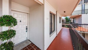 Un estrecho pasillo con una puerta blanca, una ventana con contraventanas y un balcón con barandilla de metal.