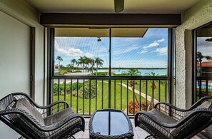 A balcony with two wicker chairs and a small table, overlooking a view of a water body and palm trees.