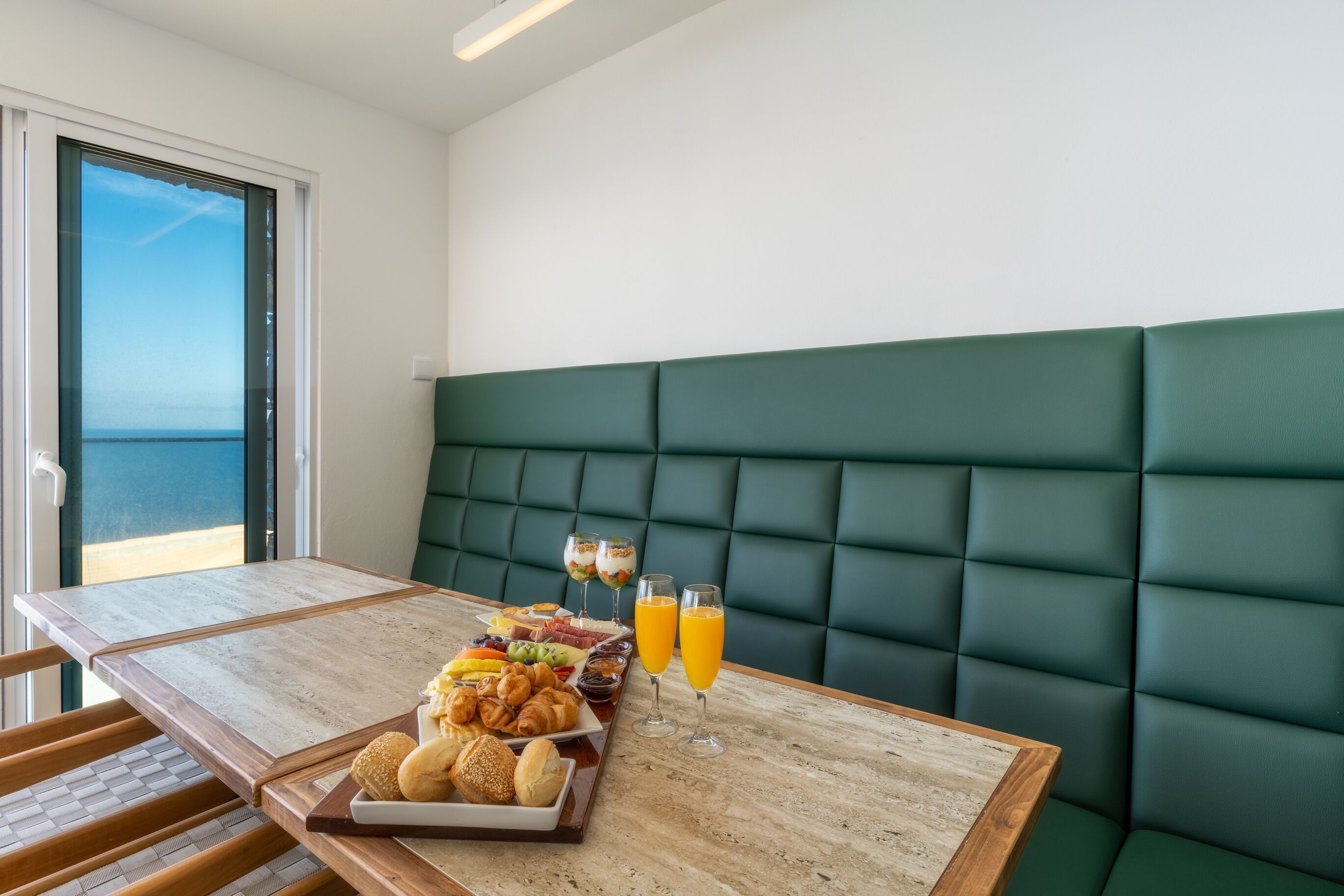 Uma área de refeições com uma mesa posta para o café da manhã, incluindo doces, frutas e bebidas, com vista para o mar através de uma janela.