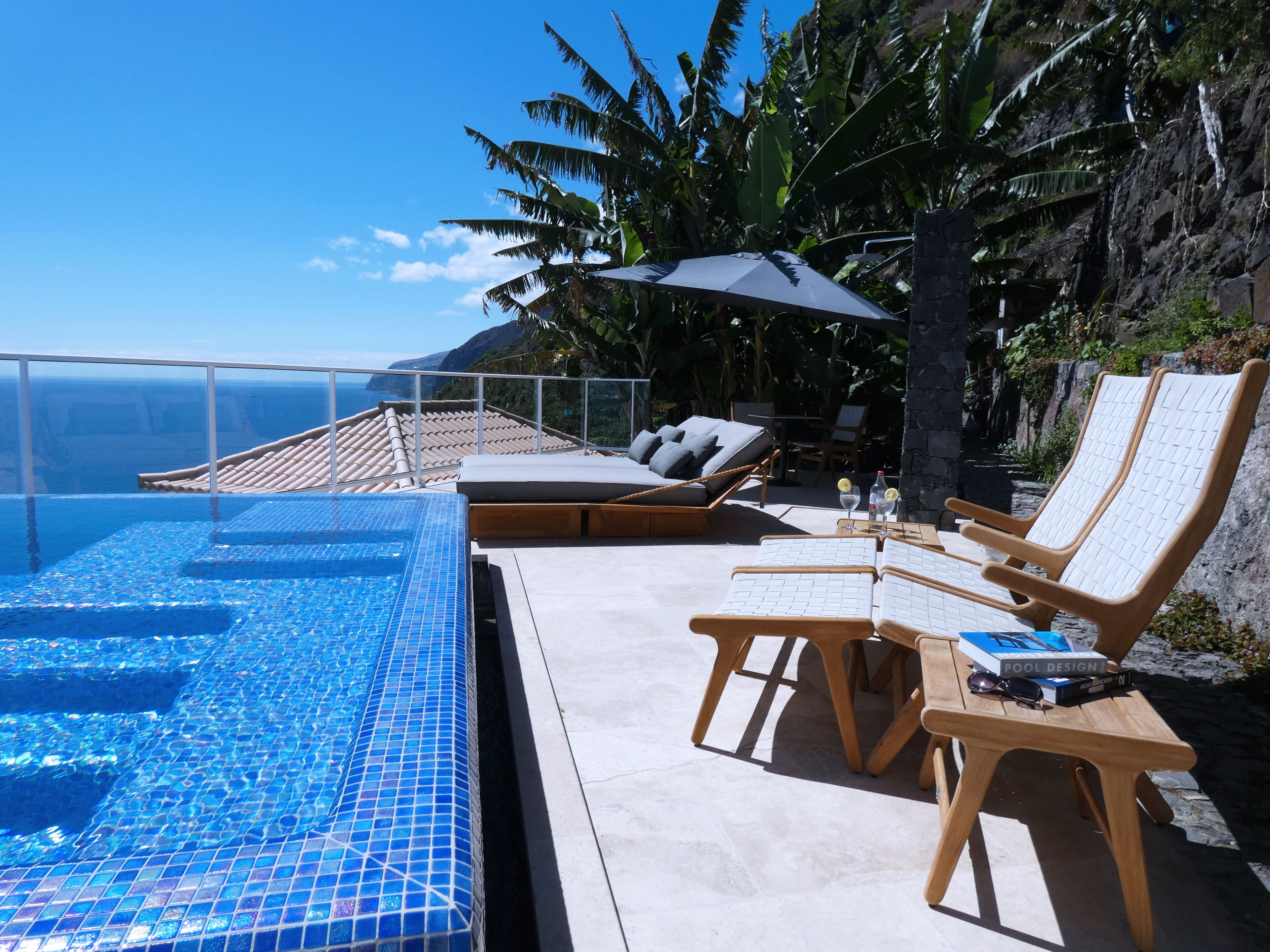 An infinity pool with clear blue tiles, wooden lounge chairs, and a view of the ocean.