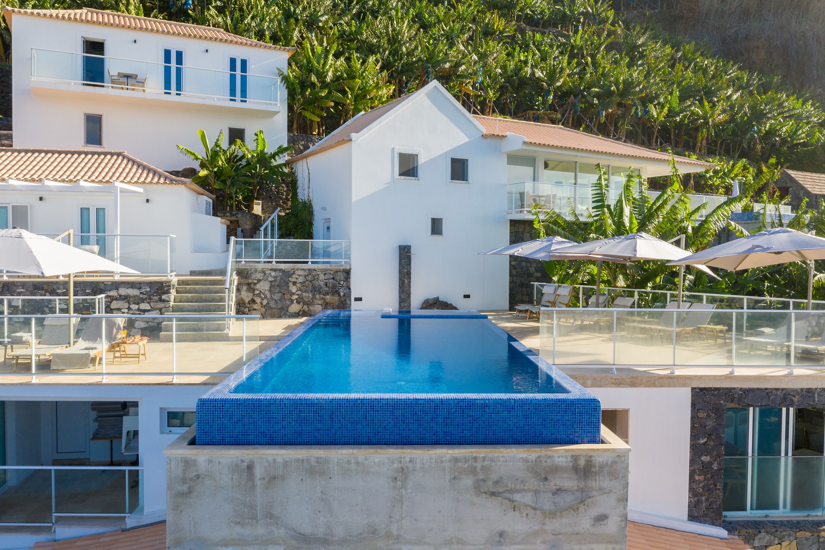 A modern pool area with a clear blue pool, surrounded by white buildings and lush greenery.