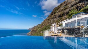 An infinity pool with a view of the ocean, a white building with a balcony, and a rocky hillside. - Escarpa - the Madeira Hideaway (Ponta do Sol)
