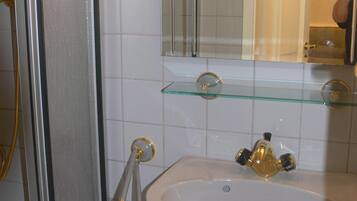 Ein Badezimmer mit einem weißen Waschbecken, einem Glasregal darüber, einer Dusche mit Glastür und einem Spiegel mit goldener Rahmung.