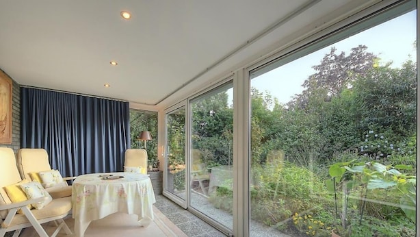 A room with a large sliding glass door leading to a garden, a round table with chairs, and a blue curtain. - Die Villa Mettmann (Mettmann)