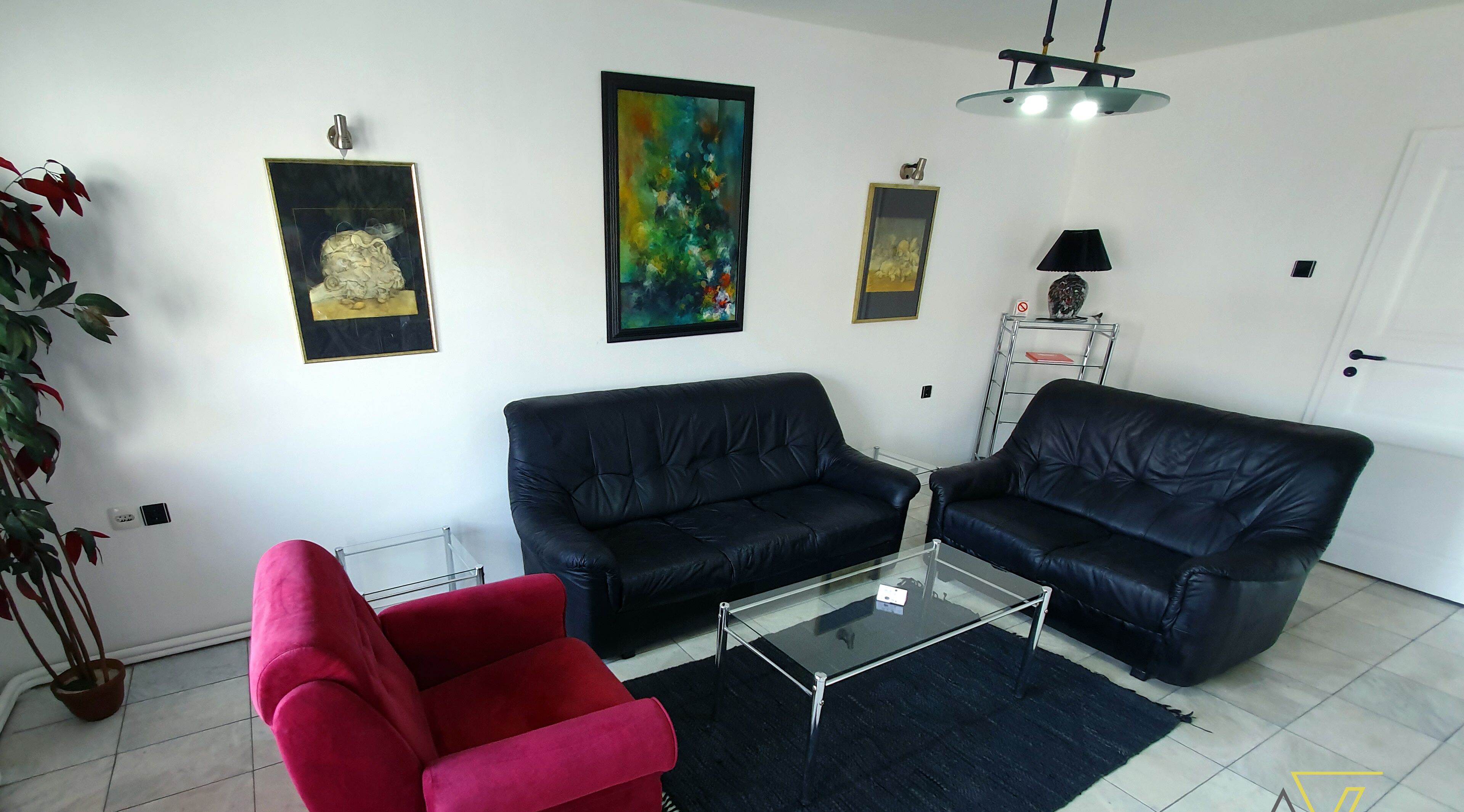 Een woonkamer met twee zwarte leren banken, een rode fauteuil, een glazen salontafel en diverse wanddecoraties.