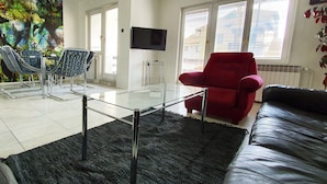 A living room with a glass coffee table, a red armchair, a black leather sofa, and a TV on the wall. - Apartments Velickovic (Soko Banja)