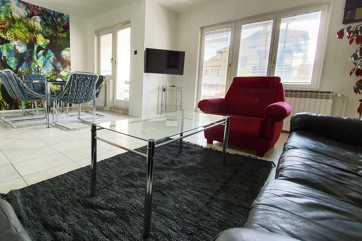 A living room with a glass coffee table, a red armchair, a black leather sofa, and a TV on the wall.