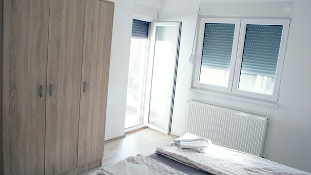 A bedroom with a wooden wardrobe, a window with blinds, and a radiator. - Apartments Velickovic (Soko Banja)