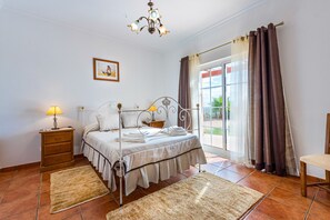 A bedroom with a four-poster bed, a nightstand with a lamp, a chair, and a view of the outdoors through a large window with curtains.