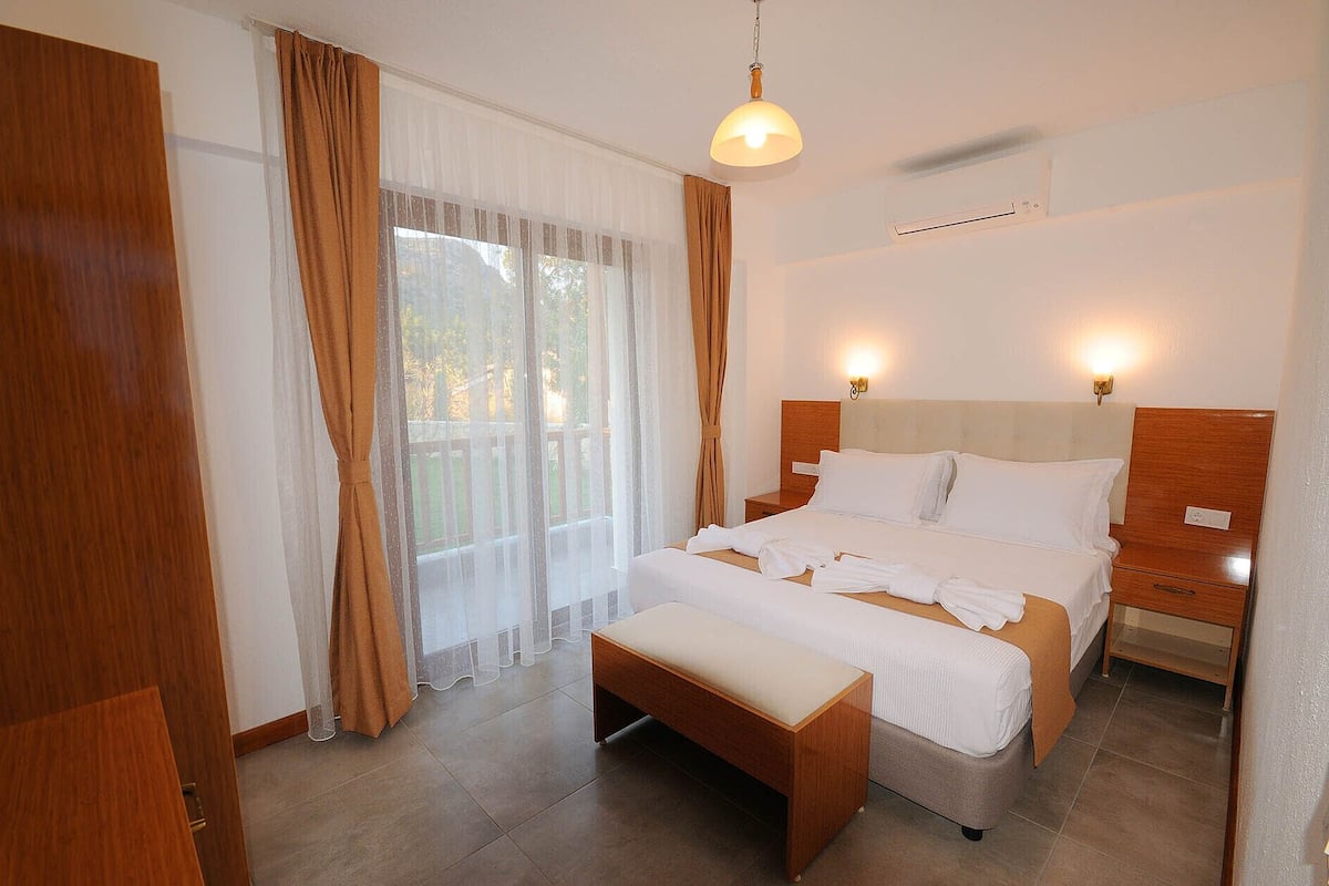 Une chambre d’hôtel avec un grand lit, une banquette, une table de chevet, une commode et une fenêtre avec des rideaux.