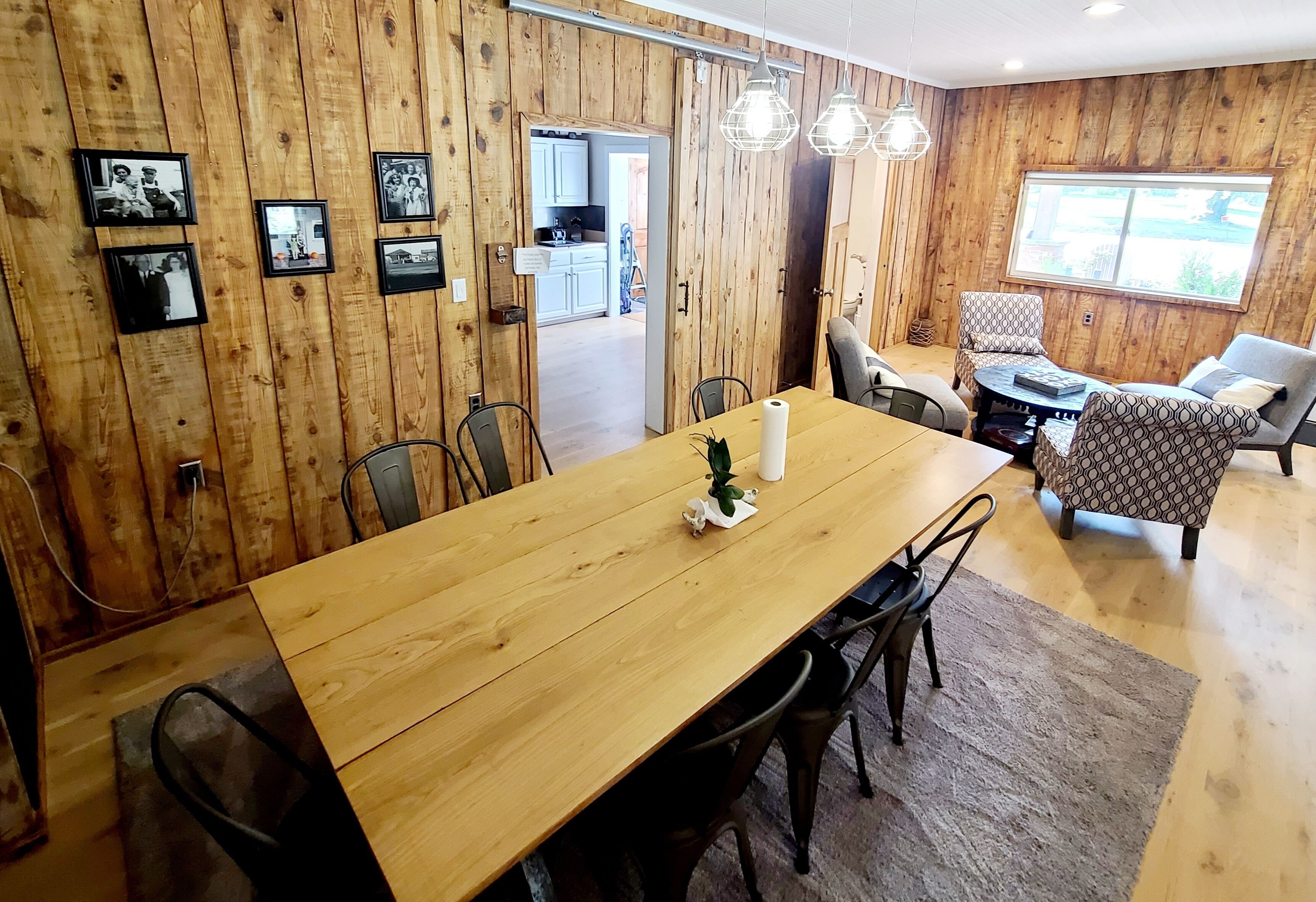 A wooden-paneled room with a dining table, chairs, and a small plant. There are framed pictures on the wall, a window, and a door leading to another room.