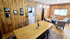 Een kamer met houten lambrisering, een eettafel, stoelen en een kleine plant. Er hangen afbeeldingen in de lijst aan de muur, en er is een raam en een deur naar een andere kamer.