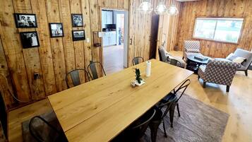 A wooden-paneled room with a dining table, chairs, and a small plant. There are framed pictures on the wall, a window, and a door leading to another room.