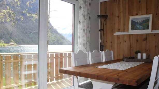 A wooden dining table with white chairs, a view of a lake and mountains through a glass door, and a wooden wall with a framed picture.