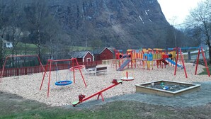 A playground with swings, a seesaw, and a slide, set against a backdrop of a mountain and a wooden fence.
