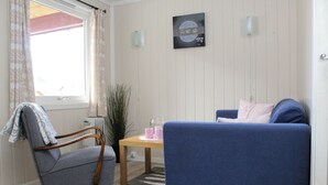 A room with a blue sofa, a wooden chair, a small table with pink items, a radiator, and a window with curtains.
