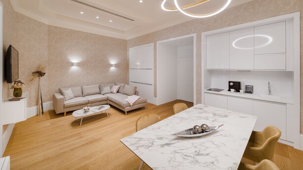 A modern living room with a sectional sofa, a dining table, and a kitchen area with white cabinetry and a built-in oven. - Hotel Vision (Budapest)