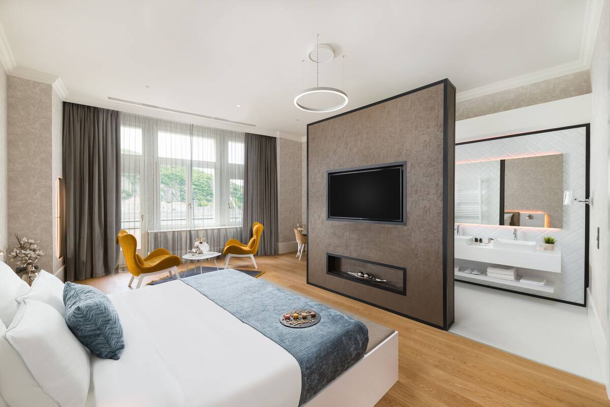 A modern hotel room with a large bed, a flat-screen TV, a built-in vanity, and a large window with curtains.