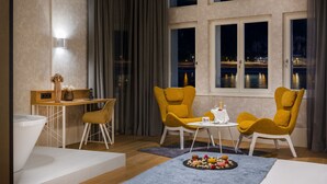 A modern hotel room with a dining area, two yellow armchairs, a small table, and a view of a cityscape at dusk. - Hotel Vision (Budapest)