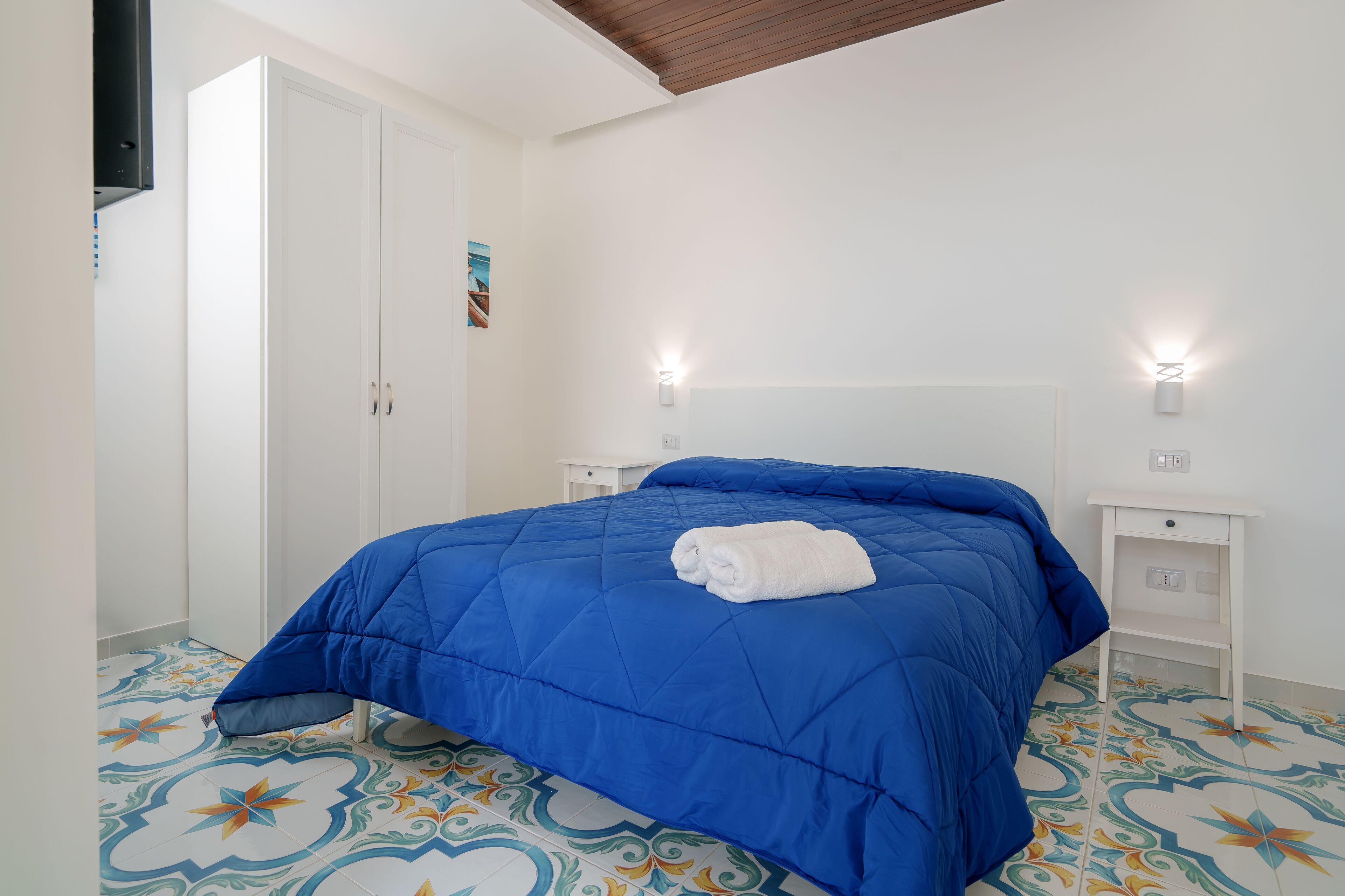 Un dormitorio con colcha azul, una puerta blanca, una mesita de noche pequeña y baldosas de suelo estampadas.