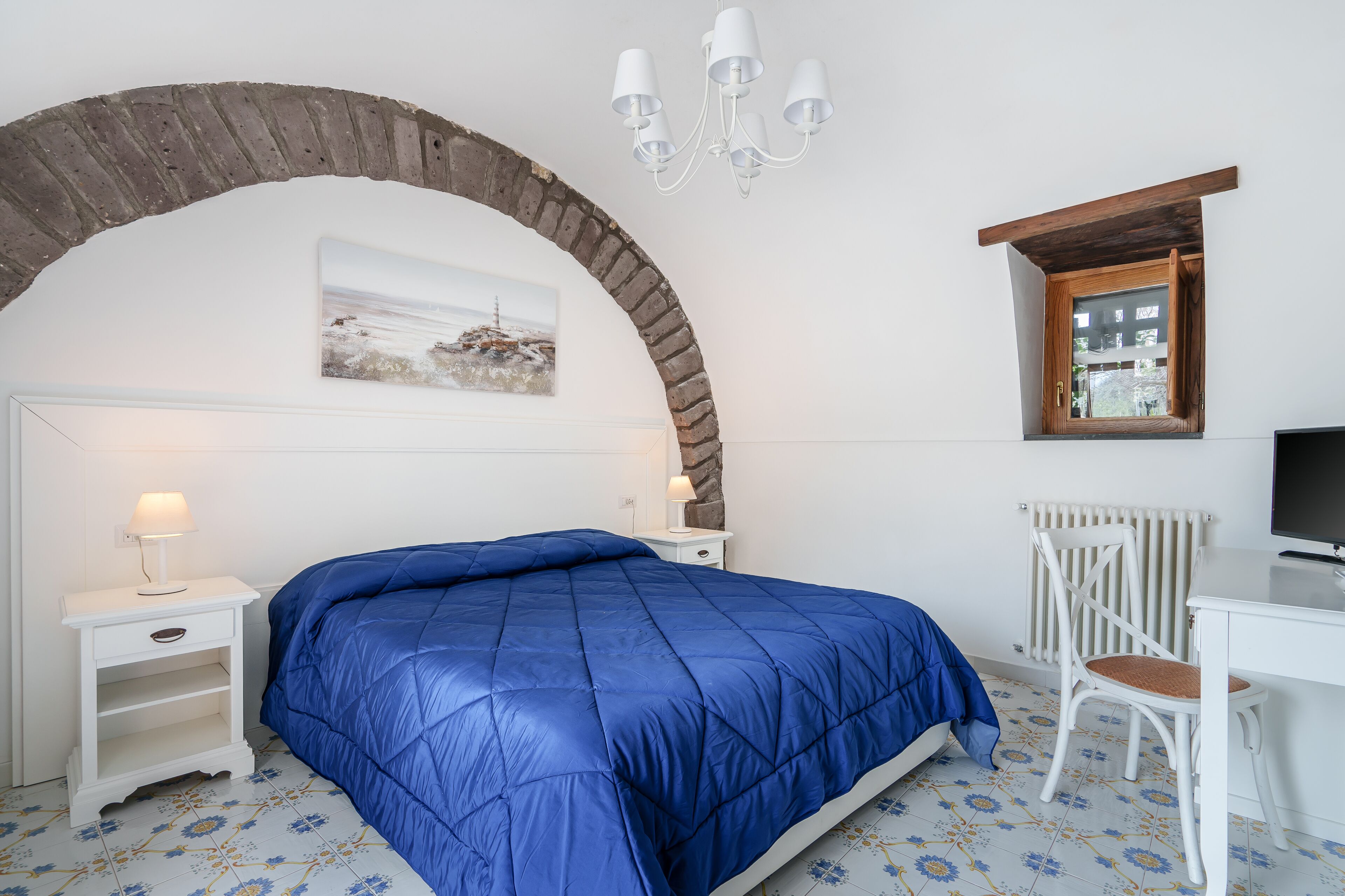 A bedroom with a bed, a desk, a chair, a lamp, a radiator, and a picture on the wall.