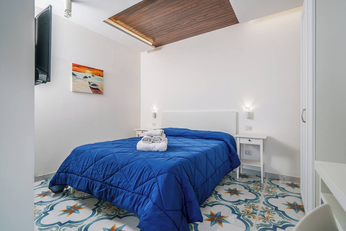 A bedroom with a blue bedspread, a painting on the wall, and a tiled floor.