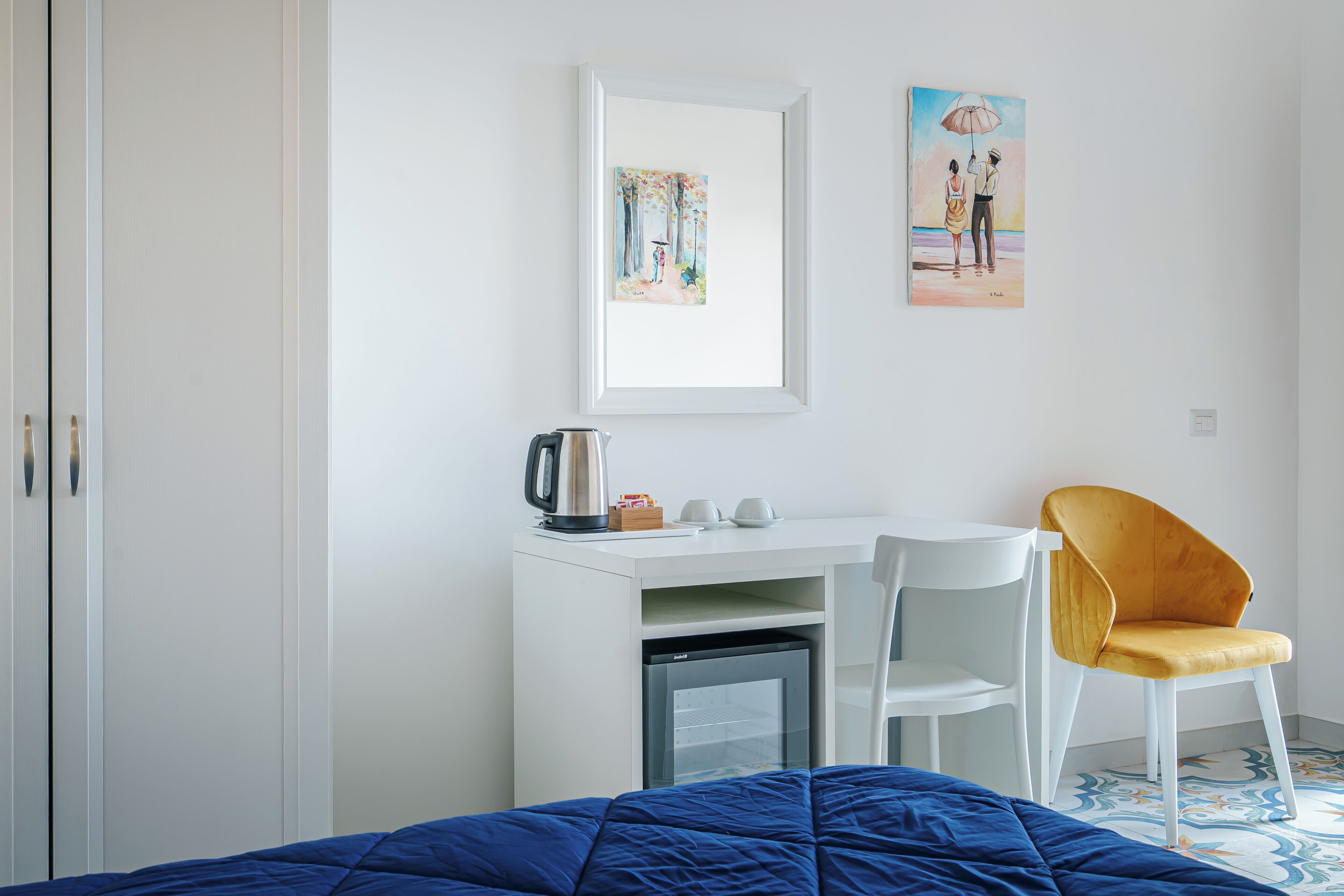 A bedroom with a bed, a desk with a chair, a refrigerator, and artwork on the wall.