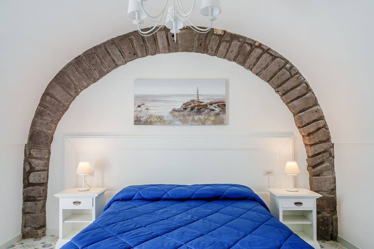A bedroom with a blue quilted bed, white nightstands with lamps, a brick archway, and a painting of a lighthouse.