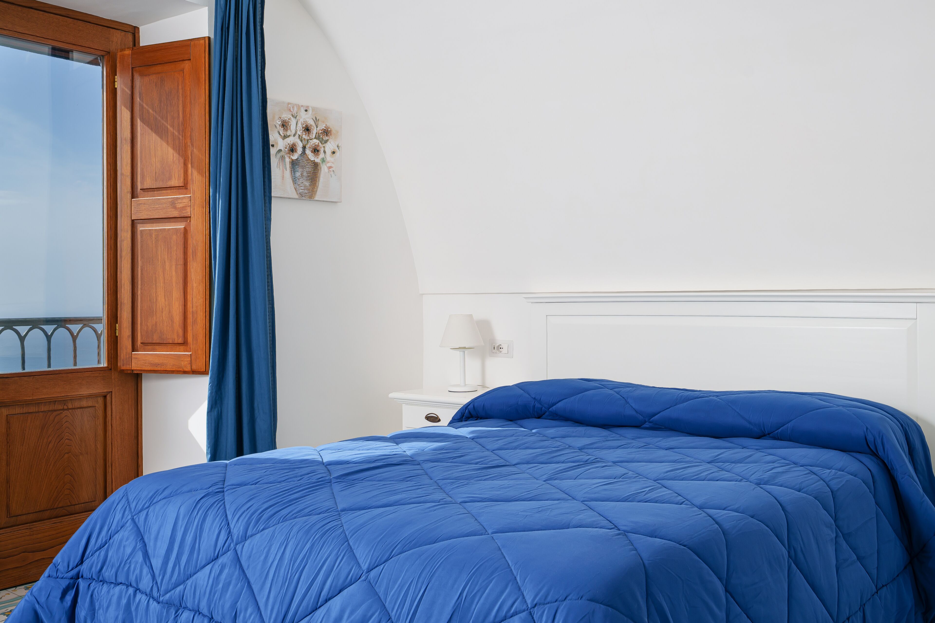 A bedroom with a blue quilted bedspread, a wooden door, a white nightstand with a lamp, and a window with a blue curtain.