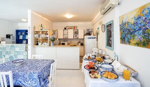 A modern kitchen with white cabinets, a dining area with a blue patterned tablecloth, and a breakfast buffet with various dishes and drinks. - Marika Studios & Apartments (Apokoronas)