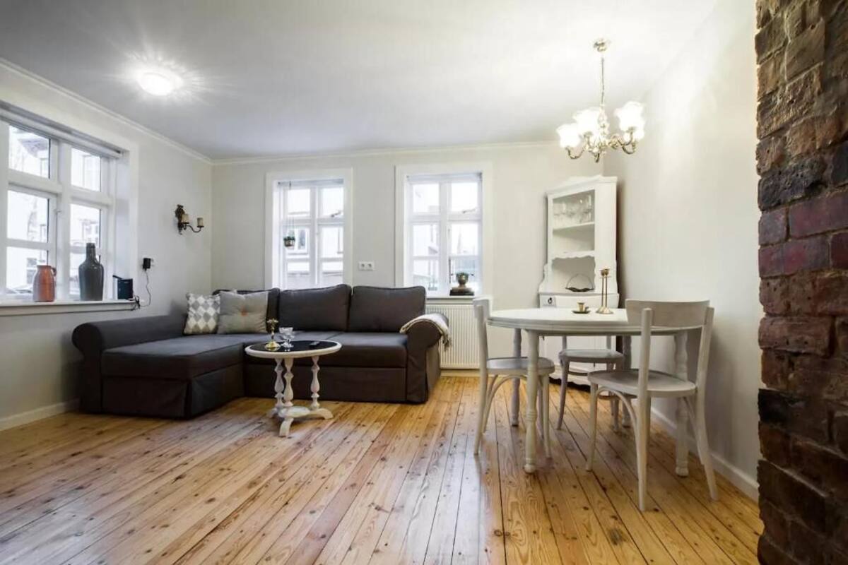 Un salon avec un canapé de couleur foncée, une table basse ronde et une table à manger avec des chaises. De grandes fenêtres aux cadres blancs, un lustre et un mur en briques complètent la pièce.