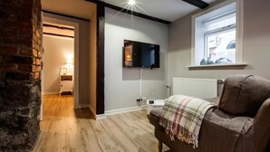 Un salon chaleureux avec un téléviseur à écran plat fixé au mur, un canapé en cuir marron agrémenté d’un plaid, et un plancher en bois.