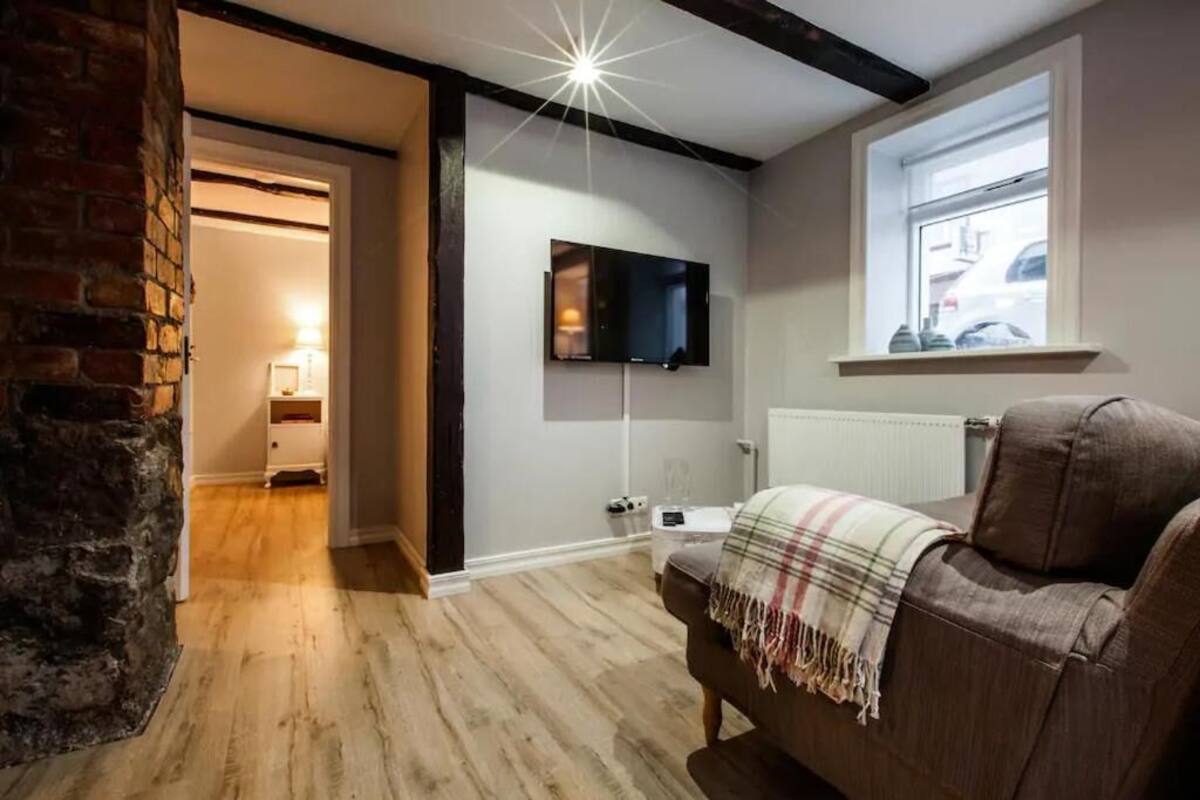 Un salon chaleureux avec un téléviseur à écran plat fixé au mur, un canapé en cuir marron agrémenté d’un plaid, et un plancher en bois.