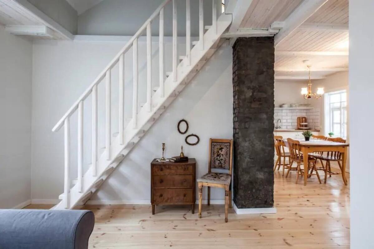 Un escalier avec des rampes blanches mène à un espace repas doté de meubles en bois, et on aperçoit une cuisine au fond.