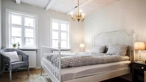 Une chambre à coucher avec un lit blanc, une chaise avec un coussin, un lustre et une table de chevet avec une lampe.