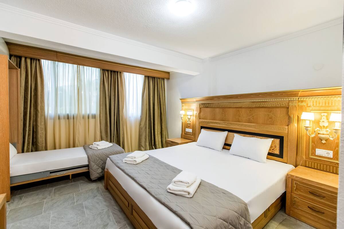 Habitación de hotel con una cama grande, una cama más pequeña, un banco, una mesita de noche y una ventana con cortinas.