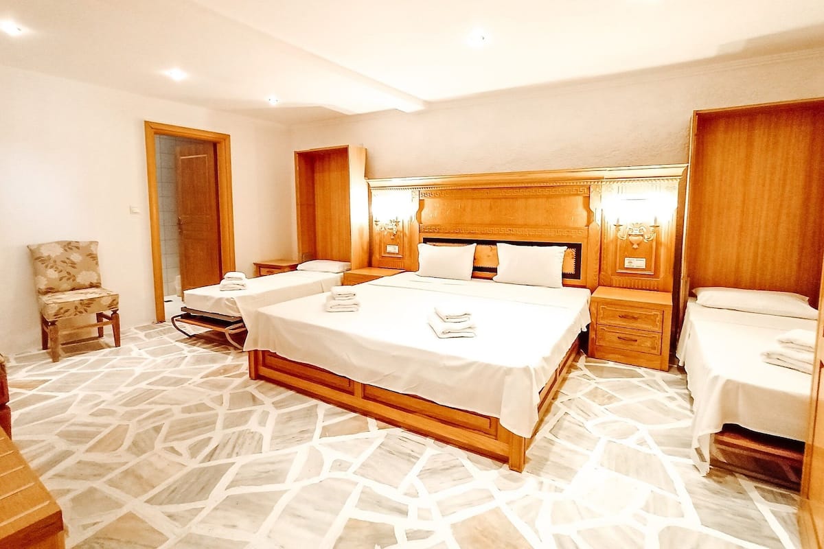 Habitación de hotel con una cama grande, dos camas más pequeñas, una silla, una mesita de noche y un cabecero de madera.