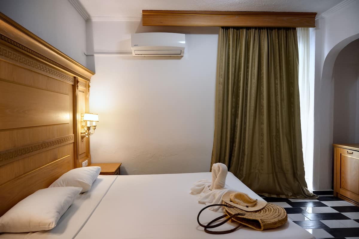 Habitación de hotel con cabecera de madera, cama con ropa blanca, una toalla y zapatillas sobre la cama, lámpara en la mesita de noche y ventana con cortinas verdes.
