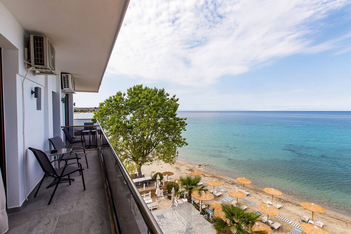 Un balcón con mobiliario exterior, con vista a una playa con sombrillas y sillas de descanso.