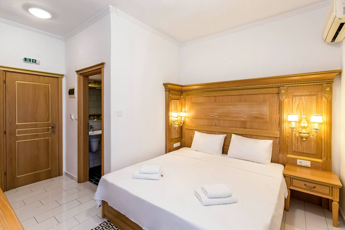 Una habitación de hotel con una cama grande, cabecera de madera, dos lámparas de noche, una mesita de noche de madera y un baño visible a través de una puerta abierta.