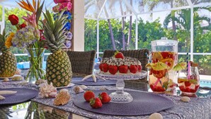 Uma área de refeições ao ar livre em ambiente tropical, com mesa de vidro, flores decorativas, uma peça central com abacaxi e um espaço para bebidas.