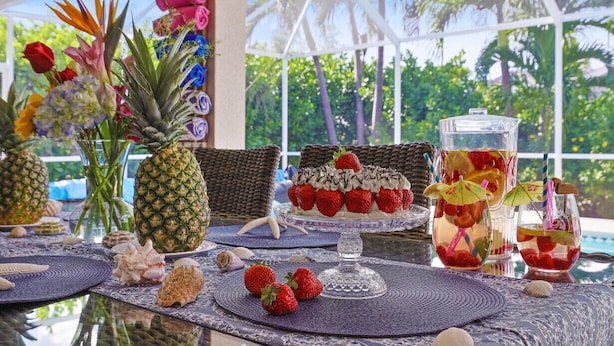 Un espace de restauration en plein air de style tropical, avec une table en verre, des fleurs décoratives, un ananas en guise de centre de table et un coin boissons.