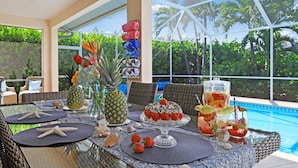 Une table à manger dressée avec un gâteau, des fruits et des boissons, donnant sur une piscine et offrant une vue sur un paysage tropical.