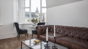 Una sala de estar moderna con un sofá de cuero marrón, una mesa de centro de vidrio y una silla negra. Una ventana ofrece vistas a edificios exteriores.